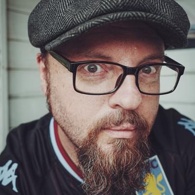 Photo of Jay Mason-Burns. He has a beard and glasses. He is wearing an Aston Villa shirt and a tweed cap.
