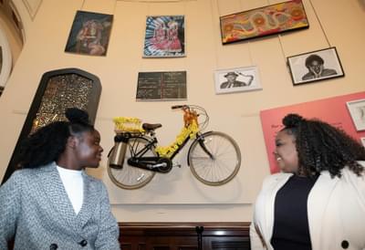 Two young women are standing either side of a bicycle that is mounted on the wall in a gallery.