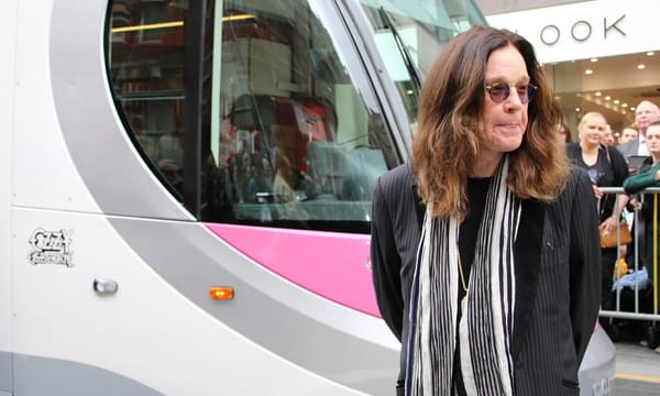 Ozzy Osbourne standing in front of a Midland Metro tram. His name is written in black and white on the side of the tram.
