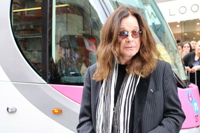 Ozzy Osbourne standing in front of a Midland Metro tram. He has long hair and has sunglasses on. He is wearing a pinstriped grey jacket and a black and white scarf.