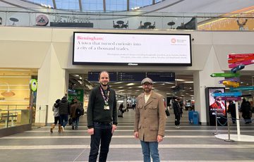 Two people standing in front of one of the screen in New Street Station.