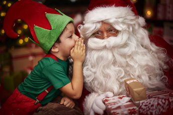A child dressed as an elf whispering into Santa's ear.