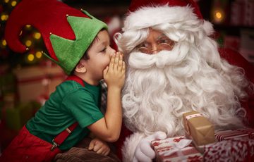A child dressed as an elf whispering into Santa's ear.