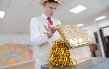 A person dressed in suit and hat opening a chest with shiny gold tinsel spilling out.