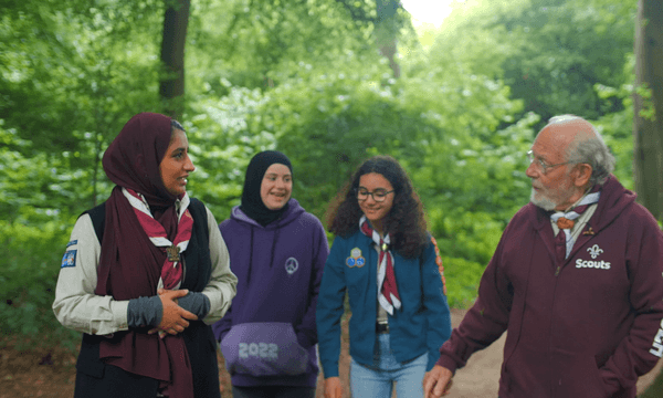 Three women and a man wearing scout uniforms are walking together and talking. They are surrounded by trees.