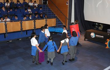 Schoolchildren hold hands in a circle performing in front of seated audience in theatre.