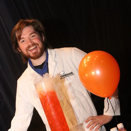 Smiling staff member in lab coat with orange balloon and tall container of bubbling red liquid.