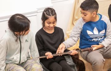 Two children sitting while holding a replica Anglo-Saxon sword with a third child looking at it.