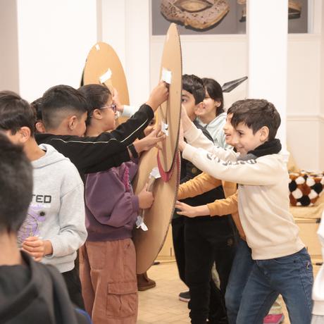 Two groups of smiling children holding cardboard shields against the other group, pretending to fight and have a battle.