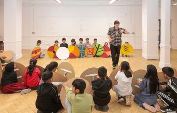 Two groups of children with cardboard shields are sitting opposite each other on the floor, a member of staff is standing.
