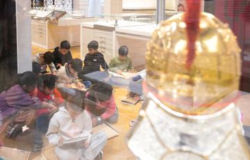 A group of children sitting on the floor of the gallery, as seen through a glass case with a helmet on display.