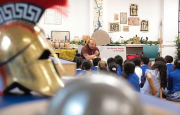 Close up of Greek Helmet on a table with children sitting listening to a staff member.