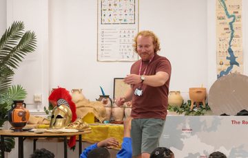 A staff member standing with a table with replica historical objects just behind. He is teaching a group of children (tops of heads are just visible).