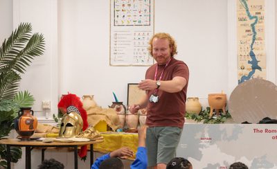 A staff member standing with a table with replica historical objects just behind. He is teaching a group of children (tops of heads are just visible).