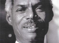 A black man looks at the camera, he's smiling. There portrait is closely cropped