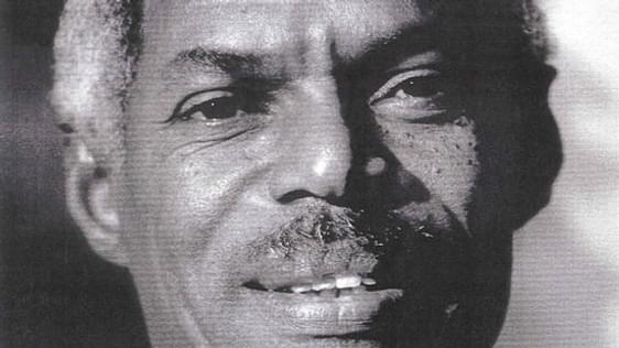 A black man looks at the camera, he's smiling. There portrait is closely cropped