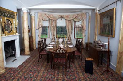 A photograph of a grand room which is very colourful and lavishly decorated. It has a large dining table and chairs, a fireplace. It has a large window with heavy curtains.