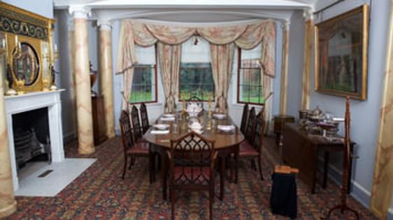 A photograph of a grand room which is very colourful and lavishly decorated. It has a large dining table and chairs, a fireplace. It has a large window with heavy curtains.