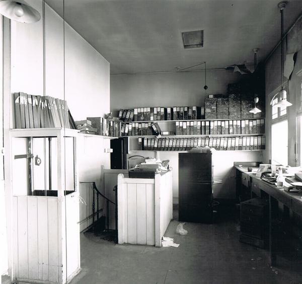 a black and white photograph of an empty office room. The walls are white, and there are lots of files on shelves.