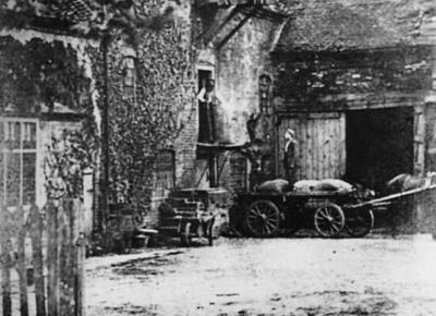 An old black and white photograph of a man standing on a cart which has been pulled by a horse. He is looking up at an opening of a large brick house