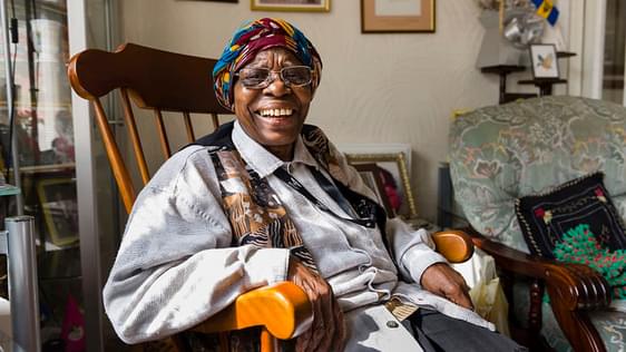 A black woman sit in a large wooden armchair looking at the camera and smiling. She's got a colourful headscarf, and pale shirt and a colourful waistcoat on.