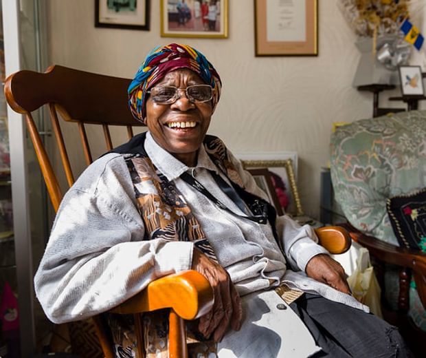 A black woman sit in a large wooden armchair looking at the camera and smiling. She's got a colourful headscarf, and pale shirt and a colourful waistcoat on.