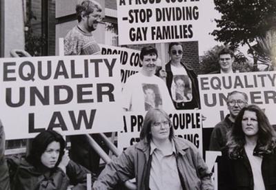 Black and white photo of protestors with signs reading "Equality under law" and "Stop dividing gay families"