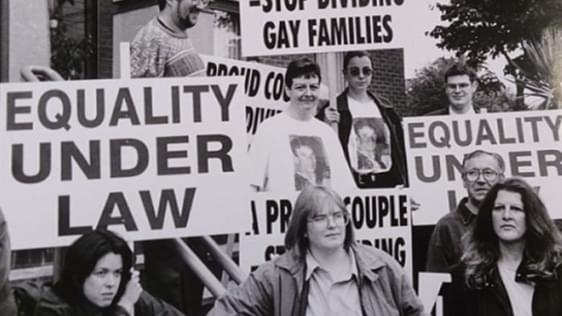 Black and white photo of protestors with signs reading "Equality under law" and "Stop dividing gay families"