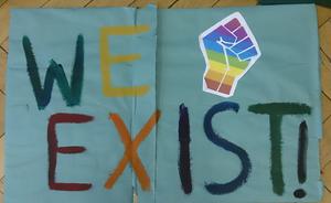 Banner with the words "We Exist" painted on it and an image of a clenched fist in rainbow colours