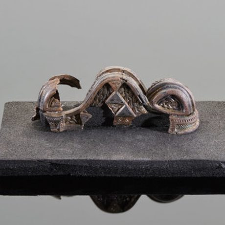 A silver object on a grey background. The object has an intricate design, but is badly damaged