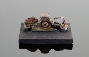 A silver object on a grey background. The object has an intricate jewel design, but is badly damaged