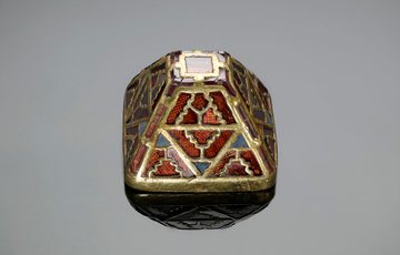 A gold and garnet pyramid, used to decorate a sword scabbard. It's has a very intricate geometric design, lots of reds and green. It's on a grey background.