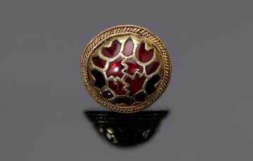 A circular gold and garnet button. It's has a very intricate geometric design, lots of reds. It's on a grey background.