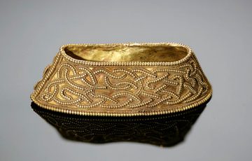 A gold object on a grey background. Gold object described as a Gold filigree hilt collar with interlace