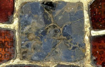 A close up photograph of glass inlayed into an object. The centre piece of glass is a blue square, and it is surrounded with smaller red squares. The red glass is textured, and almost looks like the skin of a strawberry.