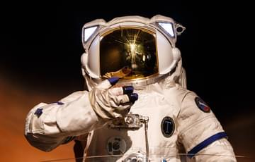 A photo of a person in a spacesuit, looking towards the camera. The photo is dark and atmospheric.