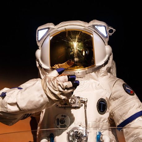 A photo of a person in a spacesuit, looking towards the camera. The photo is dark and atmospheric.