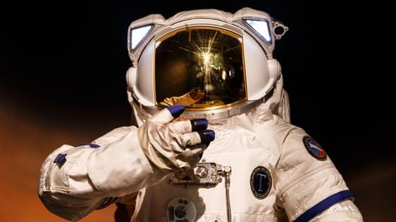 A photo of a person in a spacesuit, looking towards the camera. The photo is dark and atmospheric.