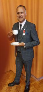 Khakan Qureshi with his British Empire Medal. He is wearing a grey suit and holding a teacup and saucer.