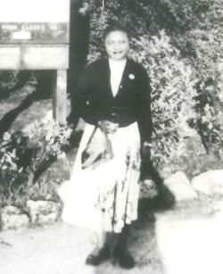 A black and white image of a black woman standing in a garden. She's wearing a dark jacked and pale skirt and carrying a bag.