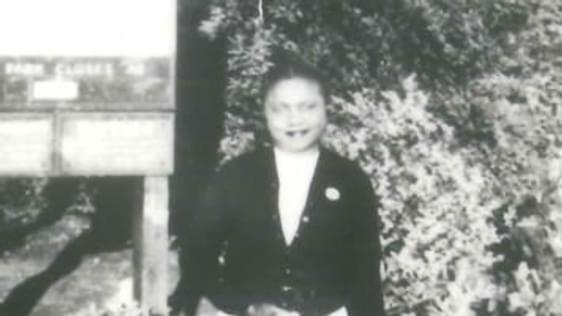 A black and white image of a black woman standing in a garden. She's wearing a dark jacked and pale skirt and carrying a bag.