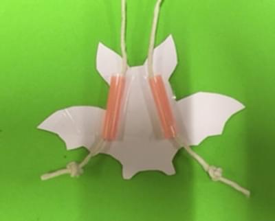 A photo of a paper bat, which has two pieces of string and straw stuck to it. It's on a green background.