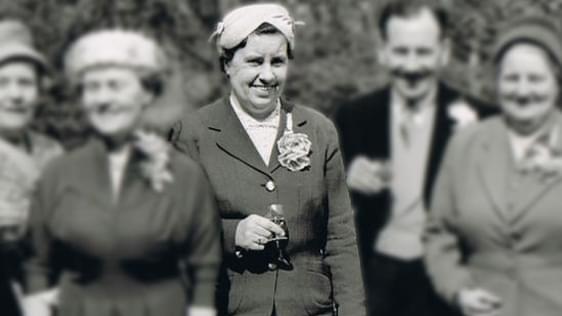An old 1940's black and white photograph of a group of people. The woman in the centre is the only person in focus. She is wearing a dark jacket with a flower on her lapel, and a hat. She is holding a glass and is looking at the camera.