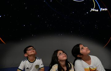 Three children in a mobile planetarium dome looking up at planets projected above.