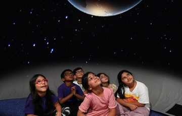 A group of children sitting in a mobile planetarium looking up at a large planet projected above.