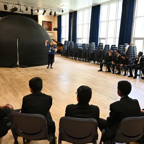 A group of secondary school pupils sat in a large circle in a school hall. A mobile planetarium with staff member is in the middle.