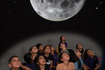 A group of children sitting in a mobile planetarium looking up at a large moon projected above.