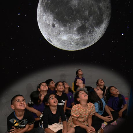 A group of children sitting in a mobile planetarium looking up at a large moon projected above.