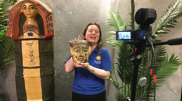 Staff member standing in front of a camera, while holding an Egyptian Mask and is surrounded by plants and a replica Egyptian coffin