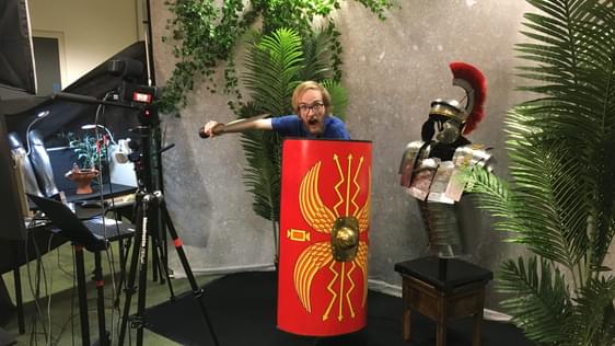 Staff member holding a replica Roman shield, sword, with a replica Roman solider costume to the side.